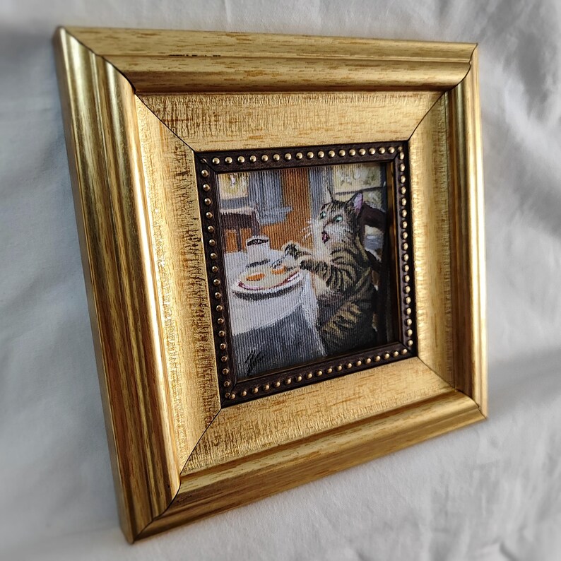 May include: A framed needlepoint artwork depicting a cat sitting at a table with a plate of food. The cat is looking at the food with a surprised expression. The frame is gold with a dark brown inner border.