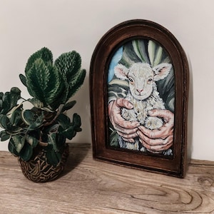 Puede incluir: Pintura enmarcada de un cordero en manos. El cordero tiene lana blanca y orejas rosas. El marco marr&oacute;n oscuro tiene forma de arco en la parte superior. A la izquierda hay una planta verde en una maceta decorativa.