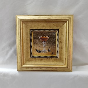 Peut inclure: Peinture encadr&eacute;e d'un cocktail dans un verre &agrave; pied, garni de cerises. L'&oelig;uvre est pos&eacute;e sur un fond dor&eacute; et encadr&eacute;e dans un cadre en bois dor&eacute;. La peinture est carr&eacute;e et repr&eacute;sente un cocktail brun.