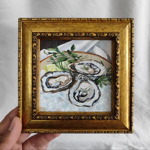 Peut inclure: Peinture encadrée de trois huîtres sur une assiette avec de la glace, des herbes et du citron. L'œuvre est dans un cadre doré orné. La peinture est de style réaliste, axée sur les thèmes culinaires.