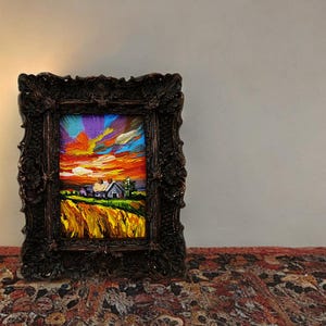 May include: A framed miniature landscape painting depicts a sunset over a field and a house. The sky is a mix of orange, purple, and blue. The ornate dark brown frame adds a vintage touch to the artwork.