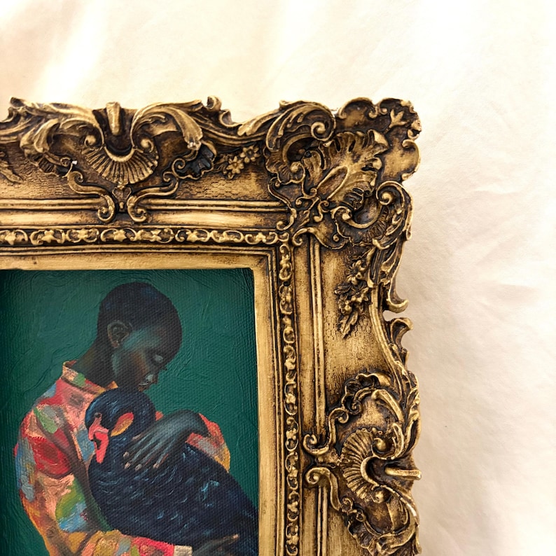 May include: An oil painting in a decorative gold frame featuring a person with dark skin wearing a colorful patterned shirt, holding a black swan against a solid dark teal background. The frame has ornate, raised floral and scroll carvings.