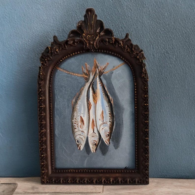 Op de afbeelding: Een ingelijst schilderij toont drie vissen die aan een touwtje hangen tegen een blauwe achtergrond. De vissen zijn geschilderd in grijstinten, bruin en wit. De sierlijke, donkerbruine lijst geeft het kunstwerk een vintage uitstraling.