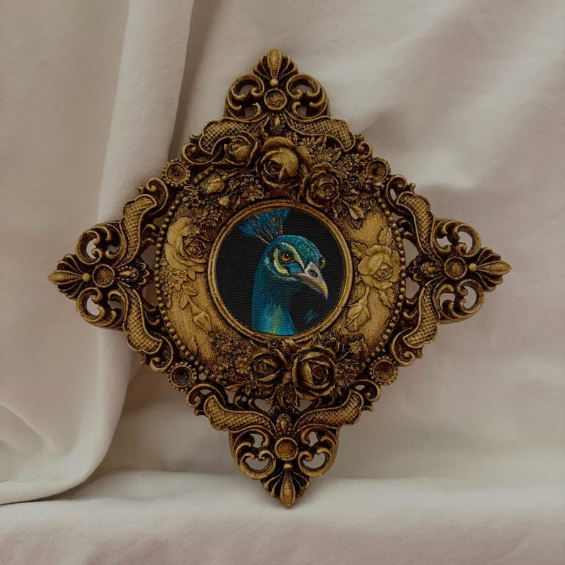 May include: A diamond-shaped, ornate gold frame featuring intricate floral and scrollwork relief details. Centered within the frame is a circular painting of a peacock head, showcasing vibrant blue feathers, a crest, and a sharp, light-colored beak.