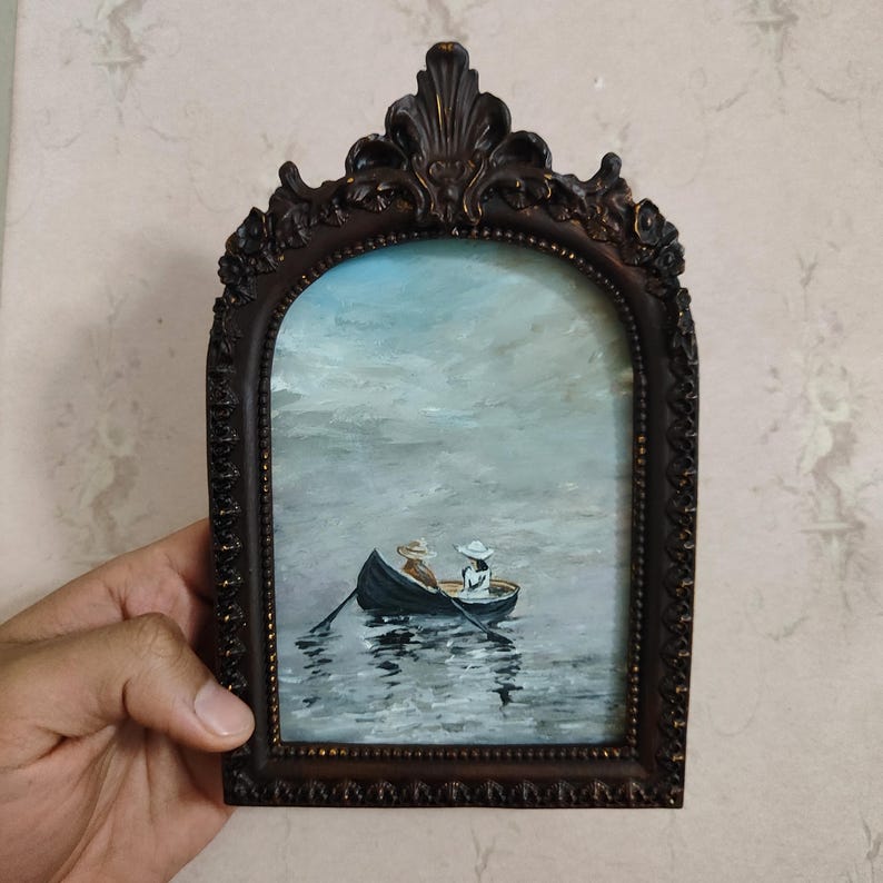 Puede incluir: Una pintura enmarcada de dos personas en un bote de remos en un lago. El cielo est&aacute; nublado y el agua est&aacute; tranquila. El marco es ornamentado y tiene un acabado marr&oacute;n oscuro.