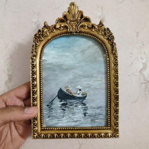 May include: A framed painting of two people in a rowboat on a lake. The painting is in a gold frame with ornate details. The sky is a light blue with white clouds. The water is a dark blue with white highlights.