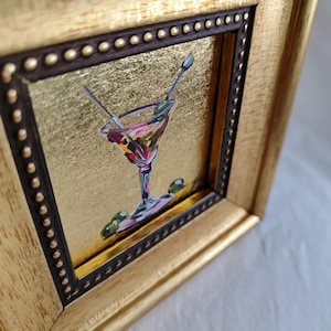 May include: A small framed painting of a martini glass with olives and a gold background.