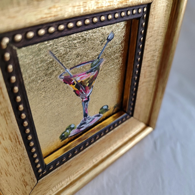 May include: A small framed painting of a martini glass with olives and a gold background.