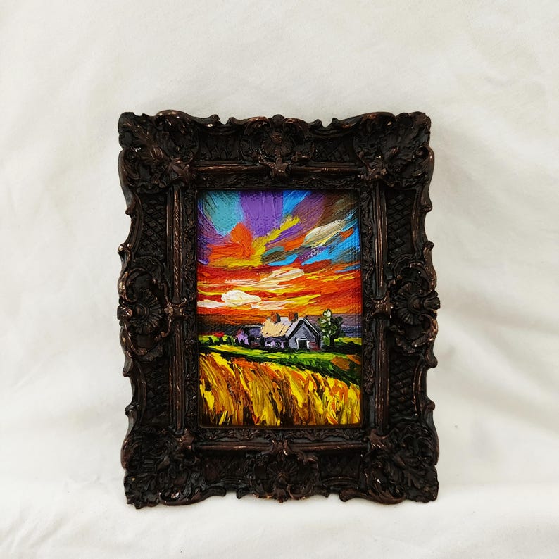 May include: A small, framed oil painting depicts a vibrant sunset over a rural landscape. The sky is a mix of orange, purple, and blue hues. A farmhouse and field of wheat are in the foreground. The ornate frame is dark brown.