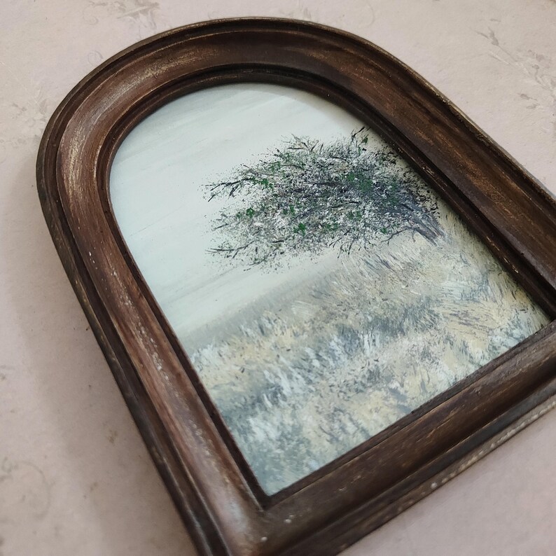 May include: An arched framed painting of a tree with green foliage in a field. The painting has a brown frame with a gold trim. The artwork depicts a landscape scene with a muted color palette.