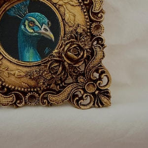 May include: A circular painting of a peacock head with blue and green feathers set inside an ornate, rectangular gold-colored frame. The frame features detailed relief carvings of roses, leaves, and scrollwork patterns along its edges.