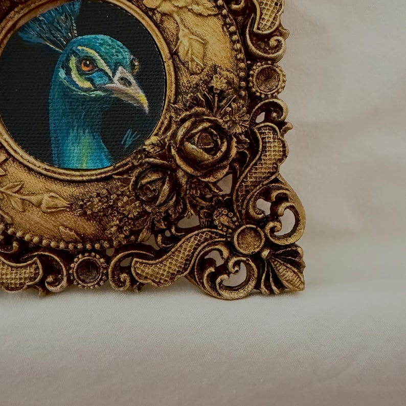 May include: A circular painting of a peacock head with blue and green feathers set inside an ornate, rectangular gold-colored frame. The frame features detailed relief carvings of roses, leaves, and scrollwork patterns along its edges.