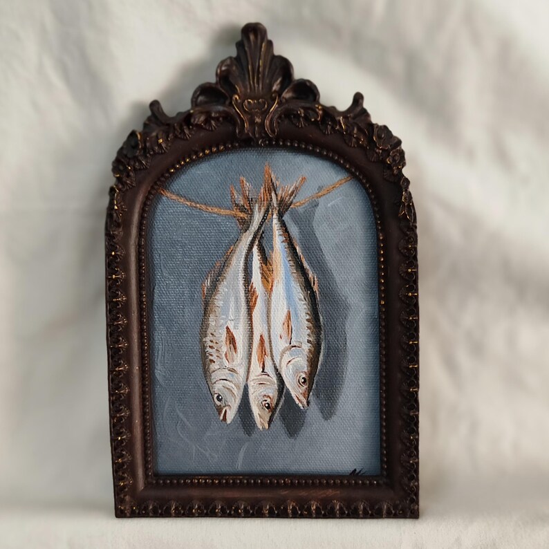 Op de afbeelding: Een olieverfschilderij van drie vissen die aan een touwtje hangen, tegen een blauwe achtergrond. Het kunstwerk is ingelijst in een donkerbruine, sierlijke lijst. De vissen hebben zilver- en bruintinten, met gedetailleerde schubben.