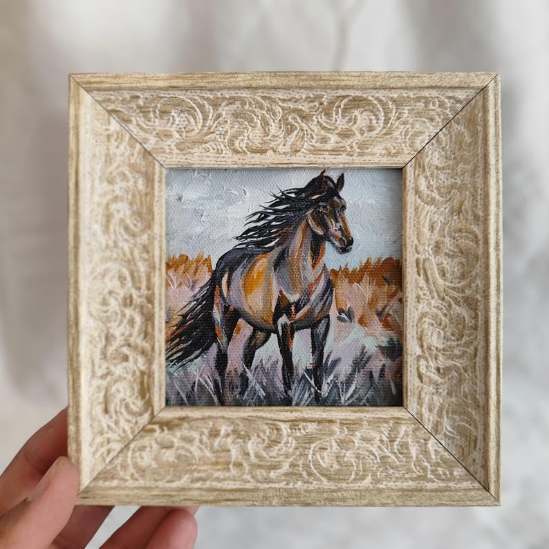 May include: A small, square framed painting of a horse. The horse is brown and tan with a flowing black mane and tail, set against a backdrop of tall grass and a cloudy sky. The frame is a light beige with ornate detailing.