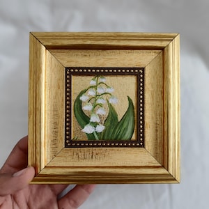 Peut inclure: Peinture encadr&eacute;e de fleurs de muguet. L'&oelig;uvre pr&eacute;sente des fleurs blanches en forme de clochettes et des feuilles vertes sur un fond dor&eacute;. La peinture est encadr&eacute;e dans un cadre en bois dor&eacute; avec une bordure int&eacute;rieure marron fonc&eacute;.