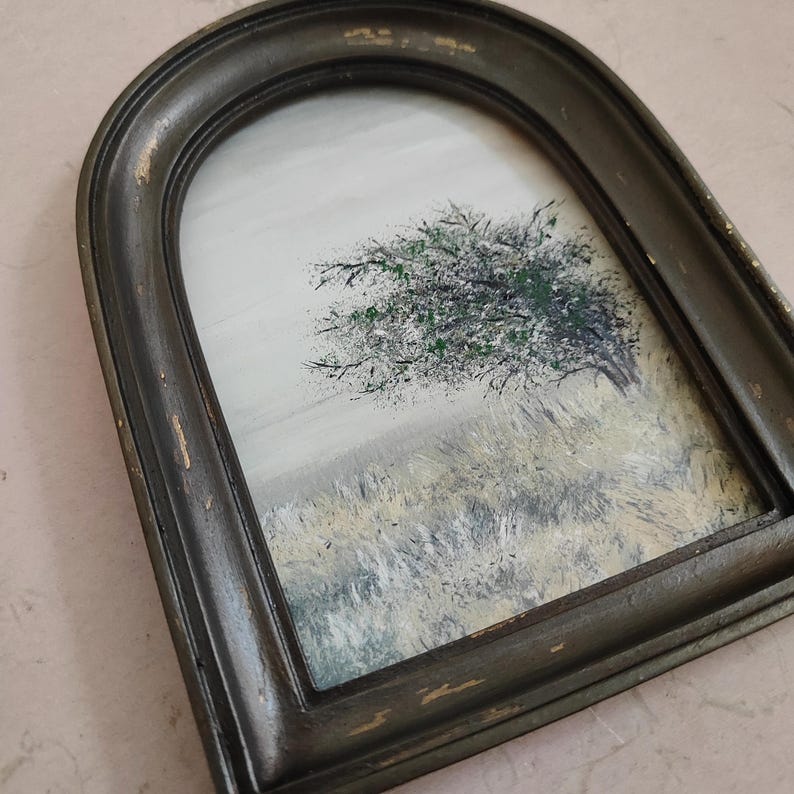May include: An arched framed painting of a tree in a field. The tree has green and black foliage. The field is painted with muted yellow and gray tones. The frame is dark with distressed edges.