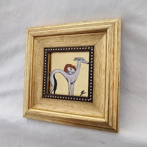 May include: A framed artwork featuring a whimsical illustration. The artwork depicts a figure with a human head and a greyhound-like body, set against a gold background. The frame is gold-colored, adding to the piece's decorative appeal.