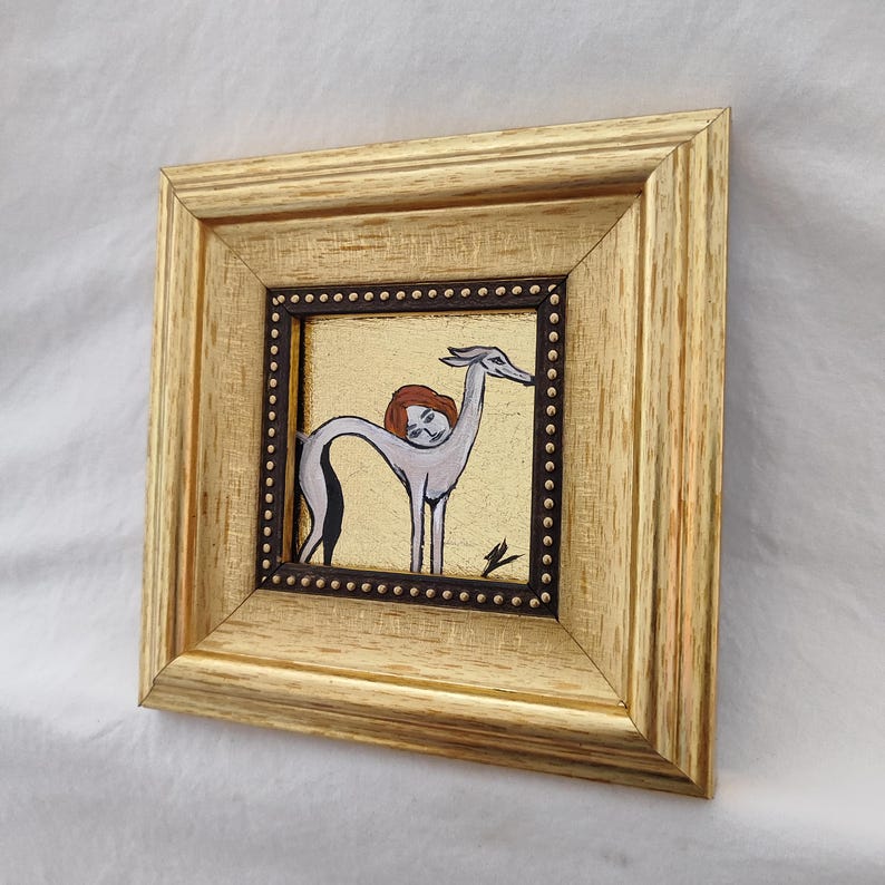May include: A framed artwork featuring a whimsical illustration. The artwork depicts a figure with a human head and a greyhound-like body, set against a gold background. The frame is gold-colored, adding to the piece's decorative appeal.