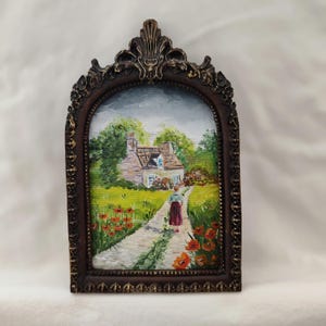 May include: A framed oil painting depicts a cottage scene with a figure walking along a path. The painting features a brown ornate frame with gold accents. The cottage has a thatched roof, surrounded by green trees and red poppies.