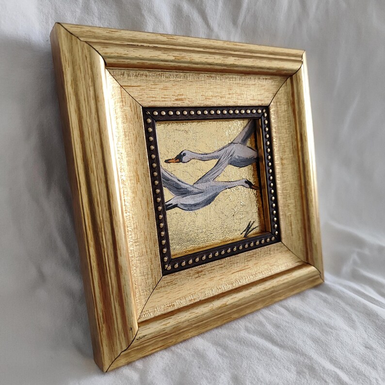 May include: A framed painting of two white swans in flight against a gold background. The frame is gold with a dark brown inner border.