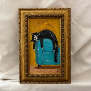 May include: A framed oil painting featuring a black cat with wide white eyes draped over a bright turquoise sofa against a mustard yellow background. The artwork is enclosed in an ornate, textured gold-colored frame with a dark brown inner border.