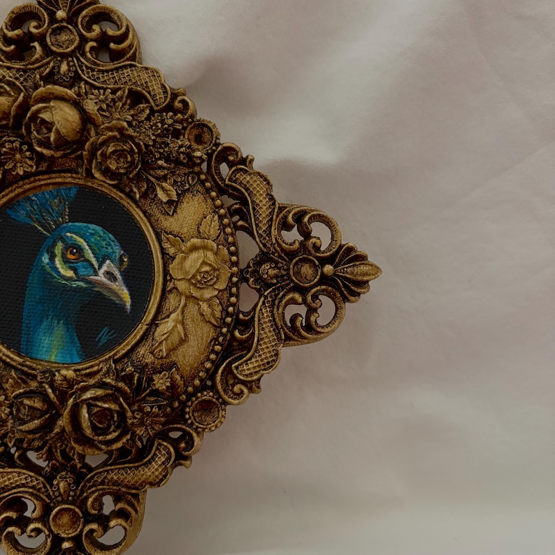 May include: A circular painting of a peacock head set within an ornate, antique gold-colored resin frame. The frame features intricate relief carvings of roses, leaves, and scrollwork, surrounding the central artwork of the blue and green bird.