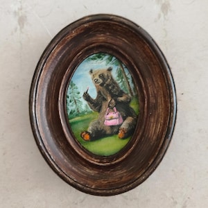 May include: A framed painting of a brown bear sitting in a forest setting. The bear is holding a pink purse and applying lipstick with its paw.