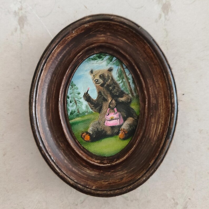 May include: A framed painting of a brown bear sitting in a forest setting. The bear is holding a pink purse and applying lipstick with its paw.