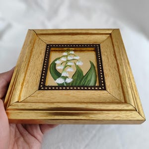 Peut inclure: Petite peinture carr&eacute;e de fleurs de muguet dans un cadre en bois dor&eacute;. L'&oelig;uvre pr&eacute;sente des fleurs blanches en forme de cloche, des feuilles vertes et un fond dor&eacute;. Le cadre a une bordure int&eacute;rieure marron fonc&eacute;.