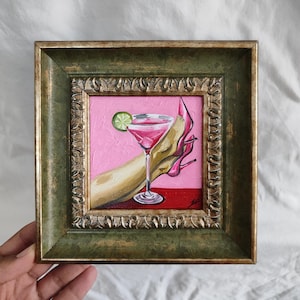 May include: A framed painting depicts a pink cocktail with a lime garnish, a hand, and a pink high heel. The artwork is set against a pink background with a red base, framed in a decorative green and gold frame.