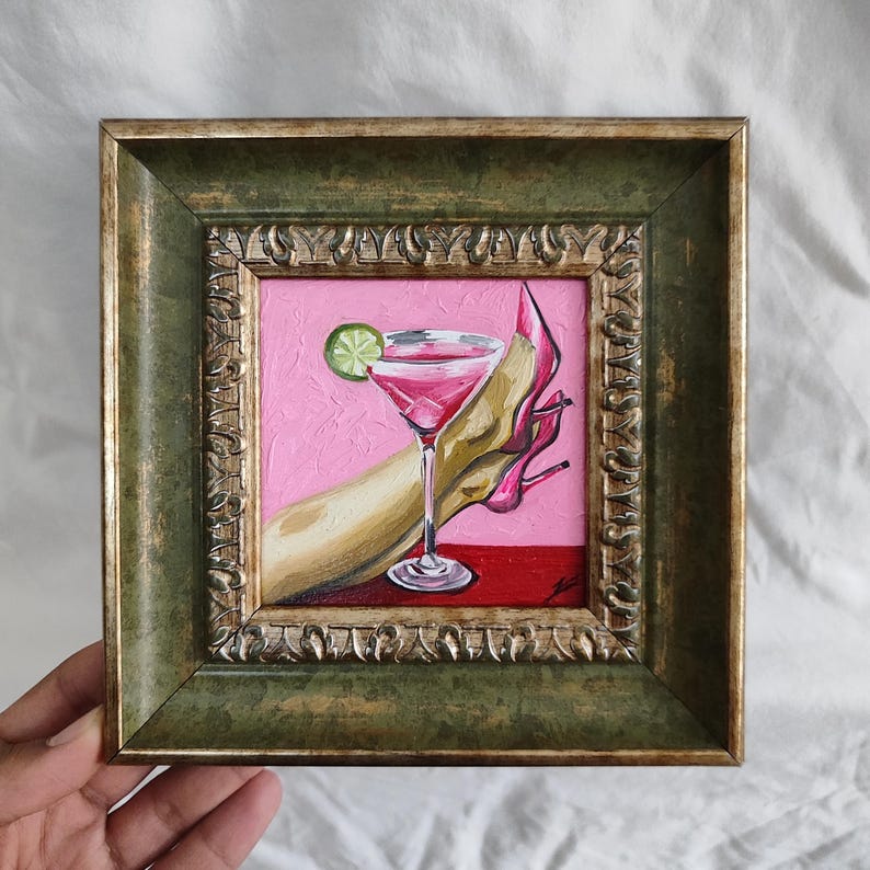 May include: A framed painting depicts a pink cocktail with a lime garnish, a hand, and a pink high heel. The artwork is set against a pink background with a red base, framed in a decorative green and gold frame.