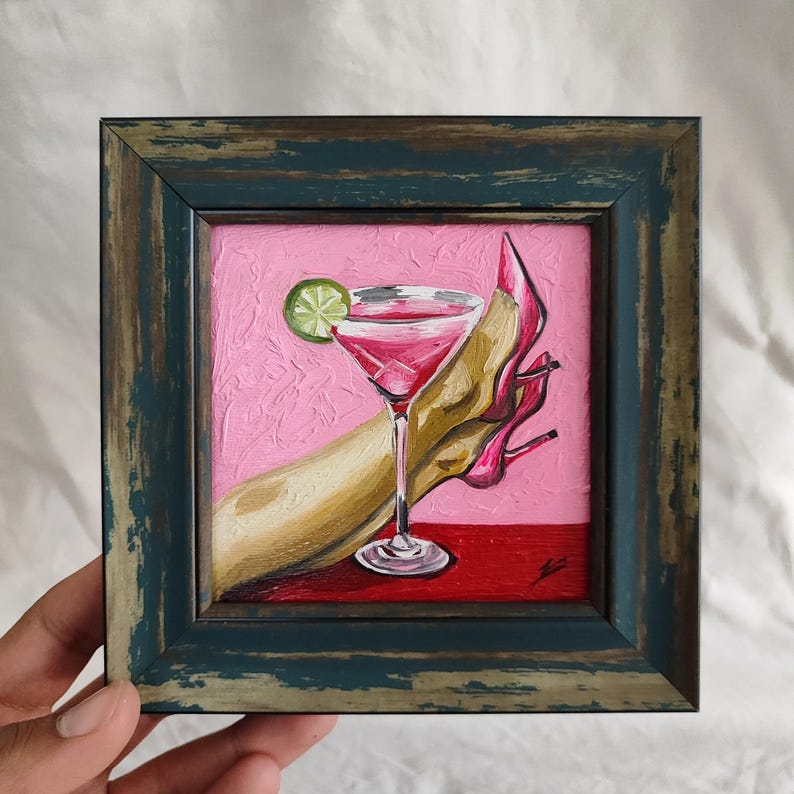 May include: A framed painting depicts a pink cocktail with a lime wedge, a high-heeled shoe, and a hand. The artwork has a pink background and a red surface. The frame is a distressed teal color.