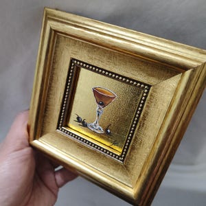 Peut inclure: Petite peinture encadr&eacute;e d'or repr&eacute;sentant un cocktail dans un verre &agrave; martini avec trois cerises noires. L'&oelig;uvre d'art a un fond dor&eacute; et est pr&eacute;sent&eacute;e en biais, soulignant la texture et les d&eacute;tails orn&eacute;s du cadre.