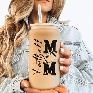 May include: A beige glass tumbler with a wooden lid and a white straw. The tumbler features the words "Football Mom" in black script and block letters, with a football graphic. The tumbler is held by a person wearing a denim jacket.