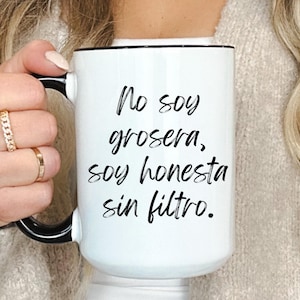 May include: White ceramic mug with a black handle and rim, featuring the Spanish text "No soy grosera, soy honesta sin filtro." The mug is held by a person wearing rings. A simple and direct design for coffee or tea.