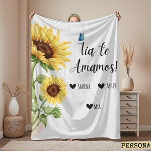May include: White blanket featuring a watercolor design of sunflowers and green leaves. The blanket has the text "Tia te Amamos!" with the names Savina, Asher, and Mia. A blue butterfly is also included.