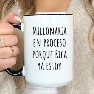 May include: White ceramic mug with a black handle and rim. The mug features the text "MILLONARIA EN PROCESO PORQUE RICA YA ESTOY" in black, a Spanish phrase meaning "Millionaire in progress because I am already rich."