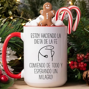 May include: A white ceramic mug with a red handle and rim, filled with marshmallows, a gingerbread man, and candy canes. The mug has text in Spanish. Festive holiday theme.