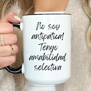 May include: White ceramic mug with a black handle and rim, featuring the text "No soy antipática! Tengo amabilidad selectiva" in black script. The mug is held by a person wearing gold rings.