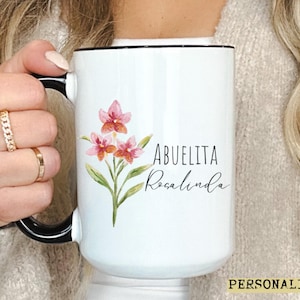 May include: White ceramic mug with a black rim and handle, featuring a floral design and the text "ABUELITA Rosalinda". The mug has a watercolor-style illustration of pink flowers and green leaves. The word "PERSONALIZADO" is at the bottom.