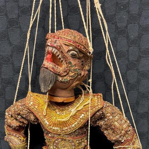 Puede incluir: Títere detallado y ornamentado con una cara feroz en forma de máscara y un elaborado disfraz. El atuendo del títere es una mezcla de oro, marrón y rojo, con intrincados detalles de lentejuelas y cuentas. Se adjuntan cuerdas para la manipulación.