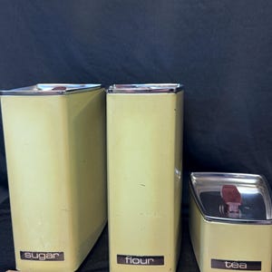 Op de afbeelding: Een set van drie vintage keukencontainers in een lichtgroene kleur. De containers zijn gelabeld met "suiker", "bloem" en "thee". Elke container heeft een zilverkleurig deksel. Een houten liniaal ligt op de voorgrond.