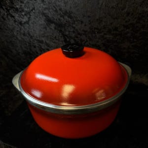 Puede incluir: Una olla holandesa de hierro fundido esmaltado de color rojo brillante con un pomo negro en la tapa. La olla tiene un borde y asas plateadas. Esta olla es ideal para cocinar a fuego lento y hornear.