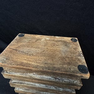 Puede incluir: Un soporte de madera escalonado, con un acabado marr&oacute;n desgastado. El soporte tiene una parte superior cuadrada y una serie de capas descendentes. Peque&ntilde;as almohadillas negras redondas est&aacute;n adheridas a las esquinas superiores.