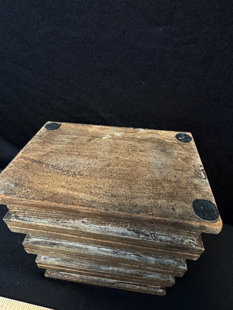 Puede incluir: Un soporte de madera escalonado, con un acabado marr&oacute;n desgastado. El soporte tiene una parte superior cuadrada y una serie de capas descendentes. Peque&ntilde;as almohadillas negras redondas est&aacute;n adheridas a las esquinas superiores.