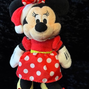 May include: A plush Minnie Mouse doll wearing a red dress with white polka dots, a matching bow, and a Santa hat. The doll has black ears, a black nose, and yellow shoes with red bows. The doll is a festive holiday item.