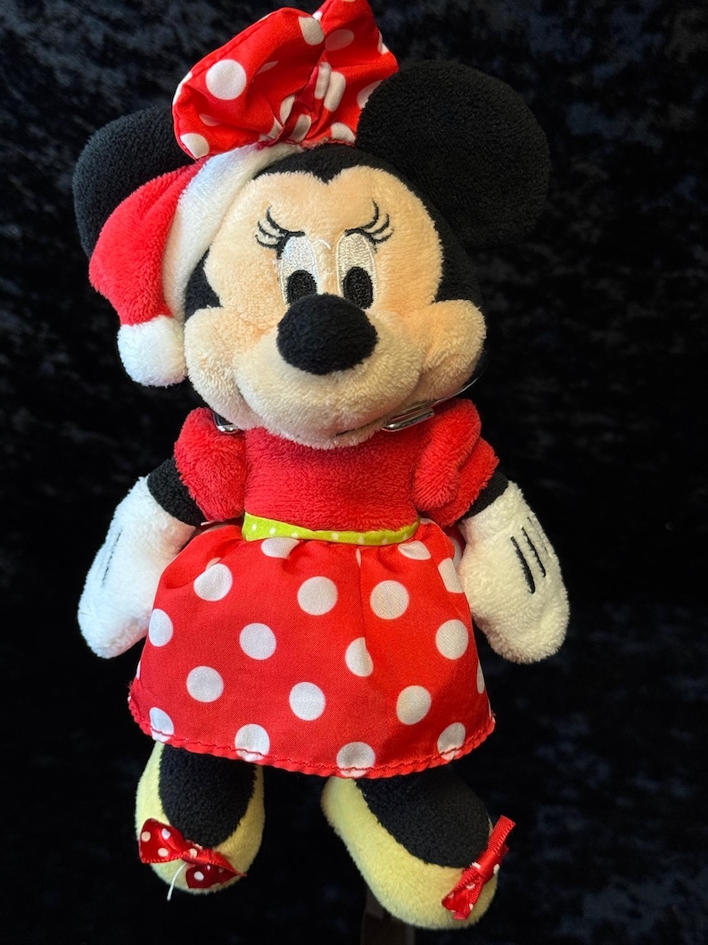 May include: A plush Minnie Mouse doll wearing a red dress with white polka dots, a matching bow, and a Santa hat. The doll has black ears, a black nose, and yellow shoes with red bows. The doll is a festive holiday item.