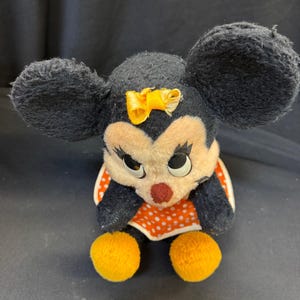 Puede incluir: Un peluche vintage de Minnie Mouse. El peluche presenta orejas negras, un lazo amarillo y un vestido de lunares rojos y blancos. El juguete tiene pies de pomp&oacute;n amarillos y una nariz roja.