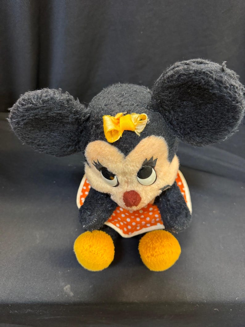Puede incluir: Un peluche vintage de Minnie Mouse. El peluche presenta orejas negras, un lazo amarillo y un vestido de lunares rojos y blancos. El juguete tiene pies de pomp&oacute;n amarillos y una nariz roja.