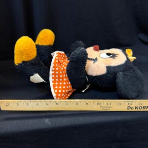Puede incluir: Un peluche vintage de Minnie Mouse, tumbado boca arriba. El peluche tiene pelo negro, pies amarillos y un vestido de lunares rojos y blancos. Se coloca una regla de madera junto al juguete para la escala.